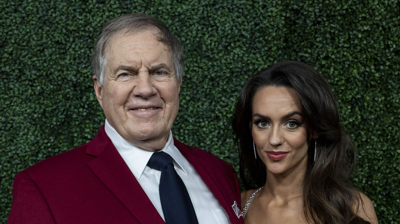 Bill Belichick posing with Jordon Hudson in front of greenery
