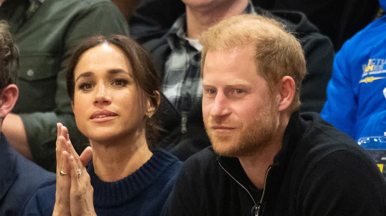Meghan Markle sitting with Prince Harry with her hands pressed together near her face.