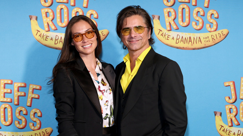 Caitlin McHugh and John Stamos smiling in matching sunglasses at the premiere of 